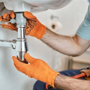 Vue en gros plan de mains portant des gants de travail orange manipulant un siphon en métal (P-trap) sous un évier blanc à Montréal.