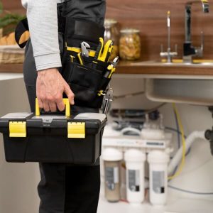 Une vue latérale d'un plombier portant une ceinture d'outils complète et tenant une boîte à outils jaune et noire, avec un système de filtration d'eau en arrière-plan à Montréal.