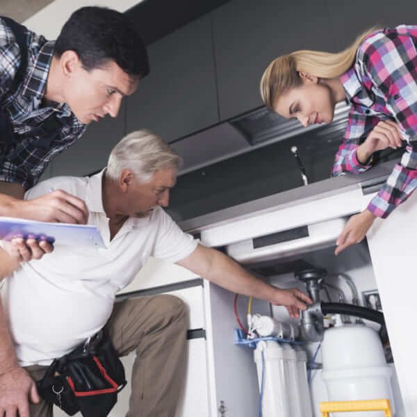 A senior plumber showing a water filtration and pipe system under a kitchen sink to a couple while holding a clipboard.