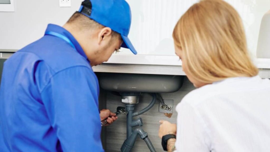 A professional plumber fixing pipes under a kitchen sink with a wrench while the homeowner watches.